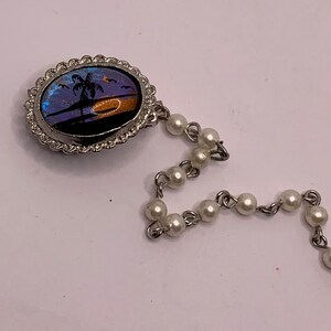 Op de afbeelding: Een zilveren armband met een ronde hanger met een vlinder vleugel ontwerp met een palmboom en zonsondergang. De hanger is bevestigd aan een ketting van witte parels.