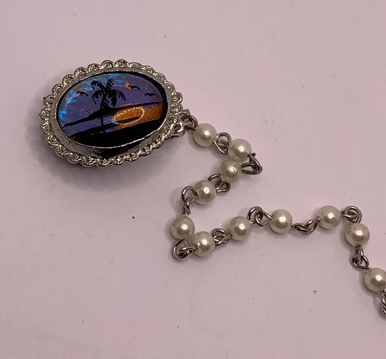 Op de afbeelding: Een zilveren armband met een ronde hanger met een vlinder vleugel ontwerp met een palmboom en zonsondergang. De hanger is bevestigd aan een ketting van witte parels.