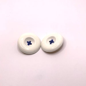 May include: Two white ceramic buttons with a blue cross stitch detail.