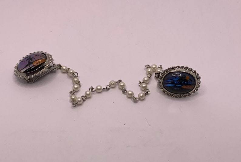 Op de afbeelding: Een zilveren ketting met witte parel kralen verbindt twee ovale broches. Elke broche heeft een kleurrijk vlinderontwerp met een zonsondergangsscene.