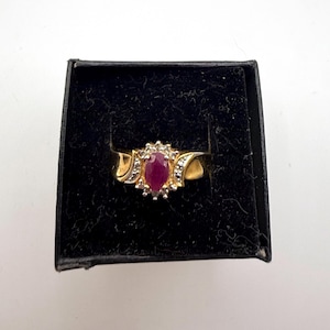 May include: A gold ring with a large, oval-shaped, red gemstone at the center, surrounded by smaller white stones. The ring is set in a black jewelry box.