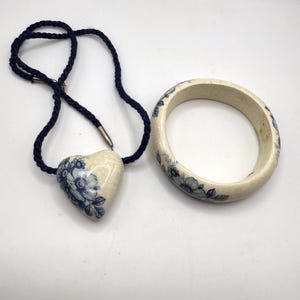 May include: A heart-shaped pendant necklace and a matching bangle bracelet. The pendant and bracelet are cream-colored with blue floral designs. The necklace has a dark blue cord.