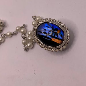 Op de afbeelding: Een zilveren ketting met witte parel kralen en een hanger met een blauwe en oranje vlinder vleugel ingelijst in een zilveren ovaal frame.