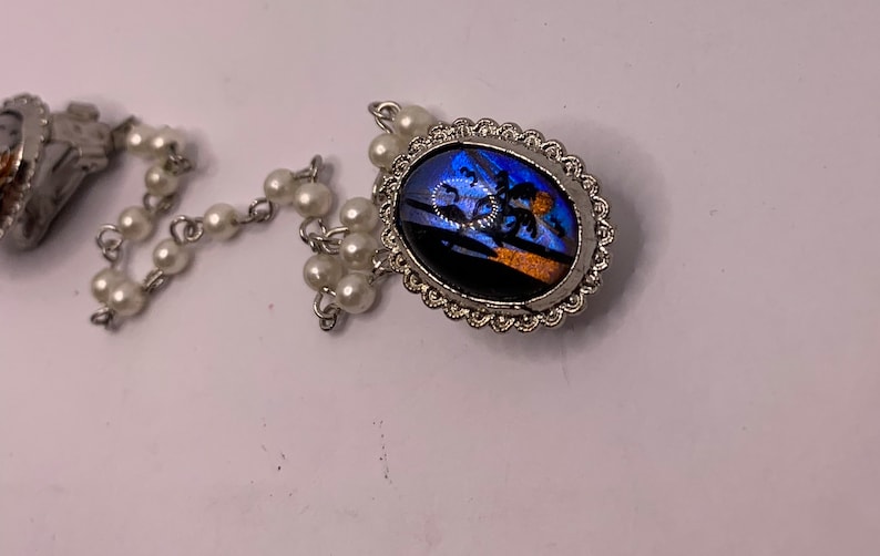 Op de afbeelding: Een zilveren ketting met witte parel kralen en een hanger met een blauwe en oranje vlinder vleugel ingelijst in een zilveren ovaal frame.