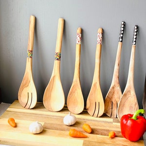 May include: Six wooden spoons with colourful beaded handles on a wooden chopping board with garlic cloves and carrots.