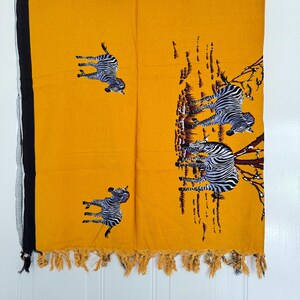 Pu&ograve; includere: Tessuto giallo senape con bordo nero e frange. Il tessuto presenta un motivo ripetuto di zebre in bianco e nero. Le zebre sono raffigurate in varie pose, alcune sembrano correre o saltare. Il tessuto &egrave; appeso su uno sfondo bianco.