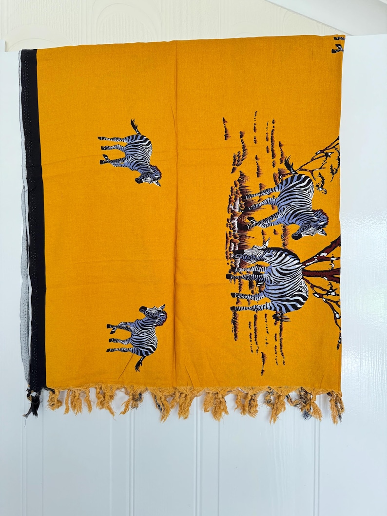 Pu&ograve; includere: Tessuto giallo senape con bordo nero e frange. Il tessuto presenta un motivo ripetuto di zebre in bianco e nero. Le zebre sono raffigurate in varie pose, alcune sembrano correre o saltare. Il tessuto &egrave; appeso su uno sfondo bianco.