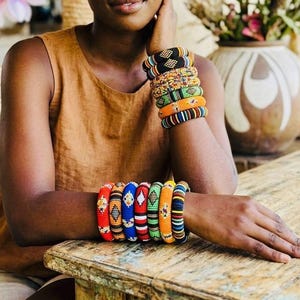 Peut inclure: Une collection de bracelets joncs perlés multicolores portés aux poignets. Les bracelets présentent des motifs géométriques complexes dans des teintes vives, notamment rouge, bleu, vert, orange, jaune, noir et blanc, faits de petites perles en verre.