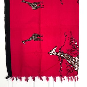 Pu&ograve; includere: Un tessuto rosso vibrante con motivi di giraffe e un bordo nero. Le giraffe hanno macchie marroni e beige. Il bordo inferiore ha nappe rosa. Probabilmente una sciarpa o uno scialle.