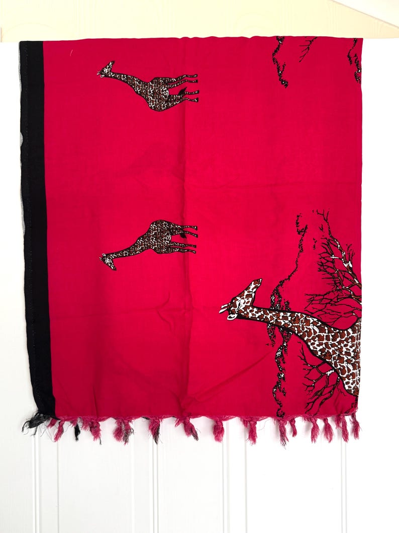 Pu&ograve; includere: Un tessuto rosso vibrante con motivi di giraffe e un bordo nero. Le giraffe hanno macchie marroni e beige. Il bordo inferiore ha nappe rosa. Probabilmente una sciarpa o uno scialle.