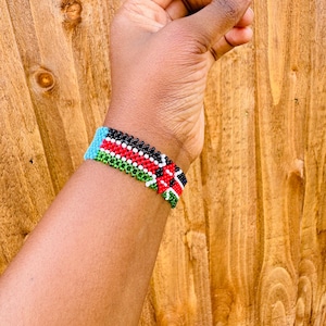 May include: A beaded bracelet featuring a repeating pattern of horizontal stripes in black, white, red, and green, with a solid light blue section at the edge. The bracelet is constructed from small, uniform spherical beads arranged in a tight grid.