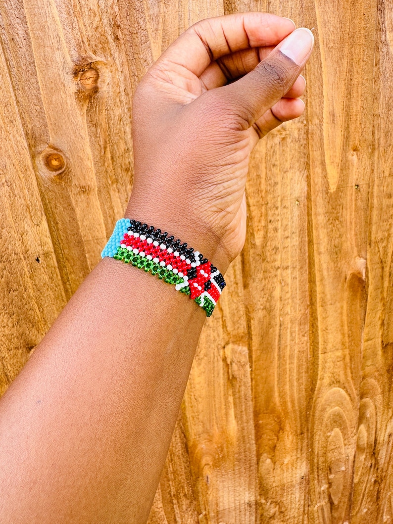 May include: A beaded bracelet featuring a repeating pattern of horizontal stripes in black, white, red, and green, with a solid light blue section at the edge. The bracelet is constructed from small, uniform spherical beads arranged in a tight grid.