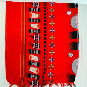 Pu&ograve; includere: Un tessuto rosso vibrante con un motivo ripetuto di tamburi stilizzati e motivi floreali. Il design include fasce colorate e un bordo sfrangiato. Il tessuto &egrave; probabilmente una sciarpa o uno scialle decorativo.