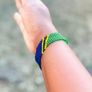 May include: A woven beaded bracelet featuring a diagonal pattern of blue, black, yellow, and green beads. The bracelet is constructed from small, uniform spherical beads arranged in a tight, grid-like structure that wraps around the wrist.