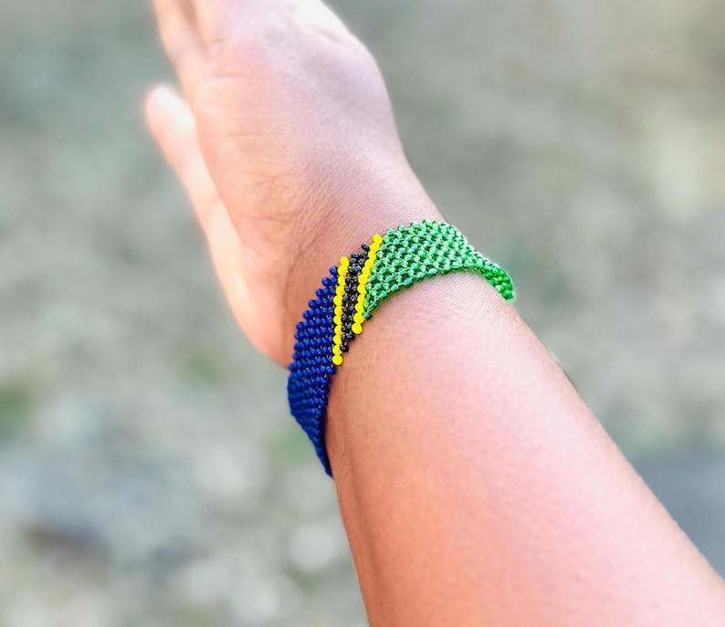 May include: A woven beaded bracelet featuring a diagonal pattern of blue, black, yellow, and green beads. The bracelet is constructed from small, uniform spherical beads arranged in a tight, grid-like structure that wraps around the wrist.