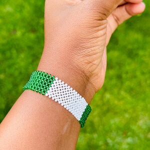 May include: A wide beaded bracelet featuring a repeating pattern of small, round beads in vibrant green and crisp white. The bracelet is constructed with a grid-like weave, showcasing a horizontal color-blocked design that wraps around the wrist.