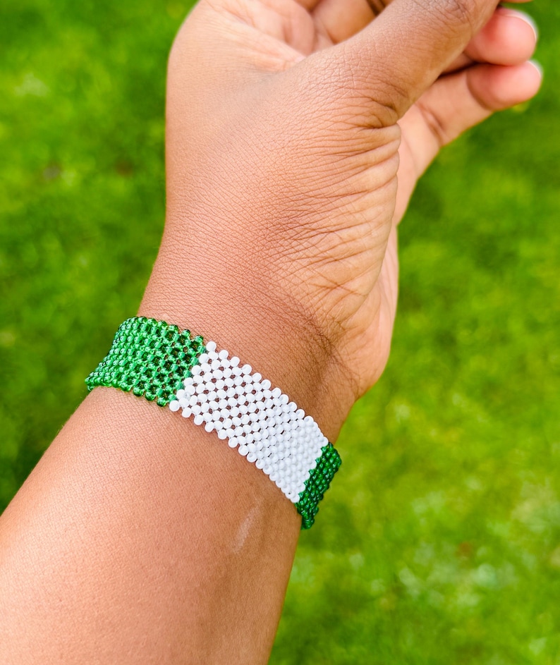 May include: A wide beaded bracelet featuring a repeating pattern of small, round beads in vibrant green and crisp white. The bracelet is constructed with a grid-like weave, showcasing a horizontal color-blocked design that wraps around the wrist.