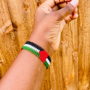 May include: A beaded bracelet featuring a repeating pattern of black, white, green, and red beads. The design consists of horizontal stripes of black, white, and green, accented by a red triangle shape on one side, creating a flag-like motif.