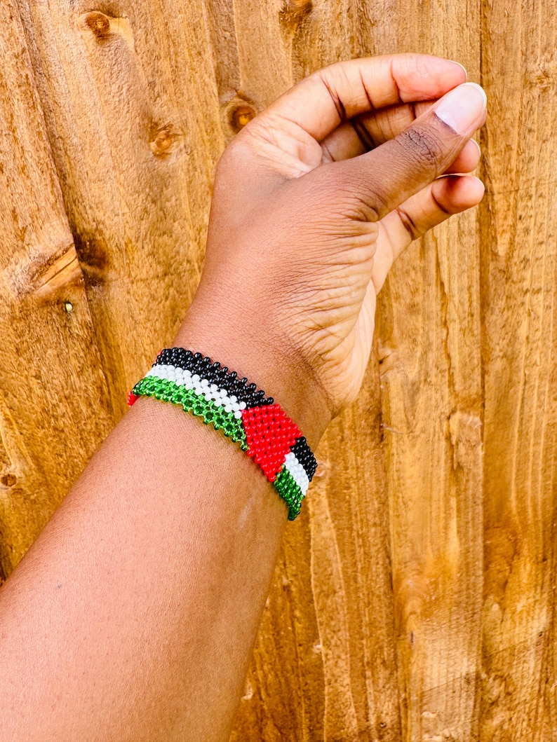 May include: A beaded bracelet featuring a repeating pattern of black, white, green, and red beads. The design consists of horizontal stripes of black, white, and green, accented by a red triangle shape on one side, creating a flag-like motif.