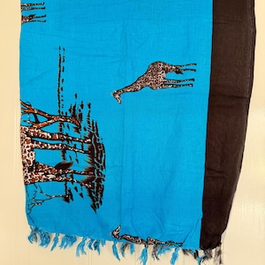 Pu&ograve; includere: Sciarpa blu con un motivo di giraffe e alberi. La sciarpa ha un bordo nero su un lato e frange blu sul fondo. Le giraffe sono marroni con un motivo a macchie. Gli alberi sono marroni con contorni neri.