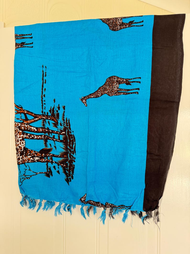 Pu&ograve; includere: Sciarpa blu con un motivo di giraffe e alberi. La sciarpa ha un bordo nero su un lato e frange blu sul fondo. Le giraffe sono marroni con un motivo a macchie. Gli alberi sono marroni con contorni neri.