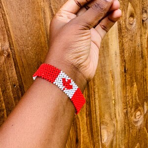 May include: A beaded bracelet featuring a repeating pattern of red and white seed beads. The design incorporates a central white square containing a red maple leaf motif, flanked by solid red sections on either side of the wristband.