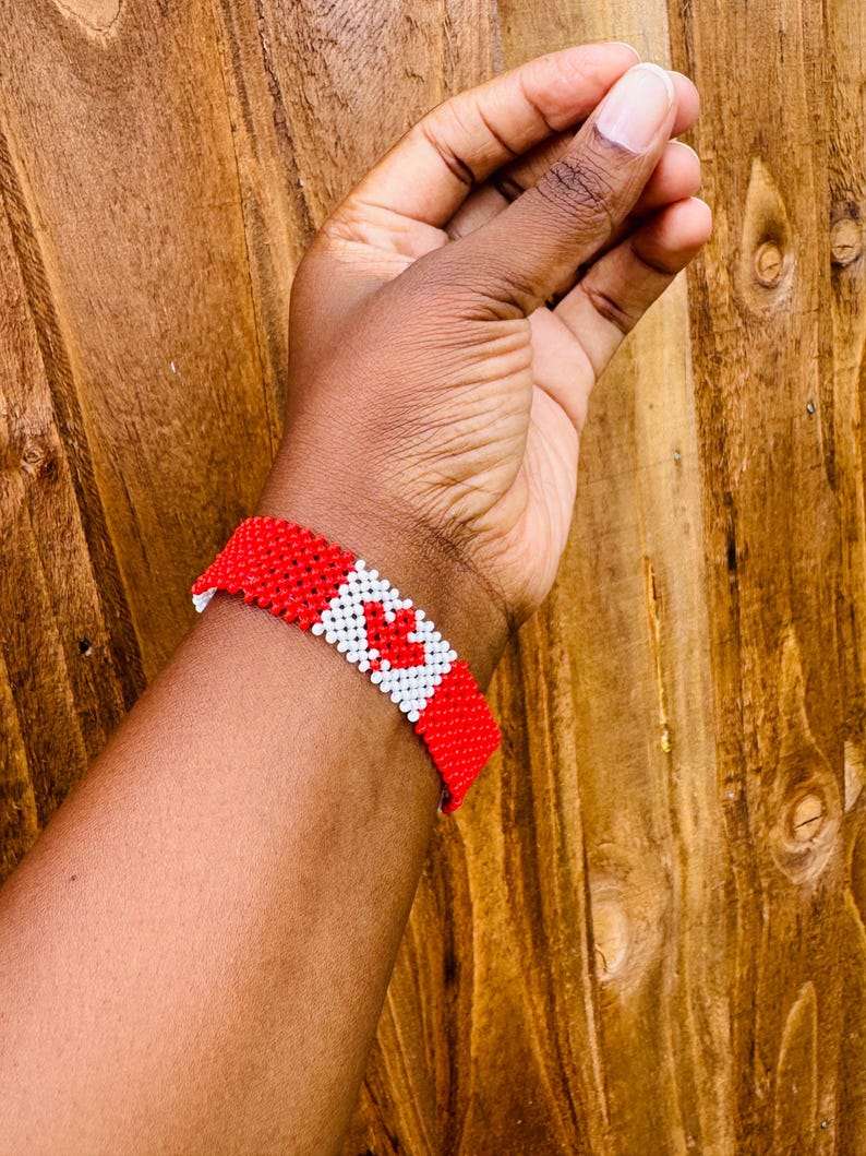 May include: A beaded bracelet featuring a repeating pattern of red and white seed beads. The design incorporates a central white square containing a red maple leaf motif, flanked by solid red sections on either side of the wristband.