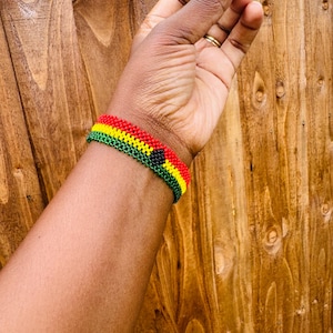 May include: A beaded bracelet featuring horizontal rows of red, yellow, and green glass beads. The design includes a small, centered black circular accent. The bracelet is constructed with a woven pattern, creating a uniform, textured surface that wraps around the wrist.