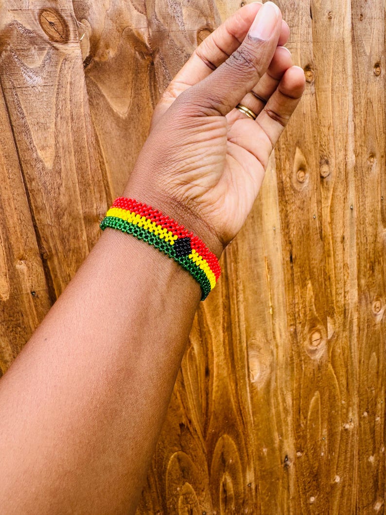 May include: A beaded bracelet featuring horizontal rows of red, yellow, and green glass beads. The design includes a small, centered black circular accent. The bracelet is constructed with a woven pattern, creating a uniform, textured surface that wraps around the wrist.