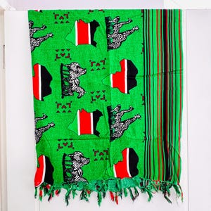 Pu&ograve; includere: Tessuto verde con un motivo che raffigura la mappa del Kenya in rosso, nero e bianco, insieme a figure di zebra e leopardo. Ha un bordo con frange e un bordo a righe rosse, nere e verdi.