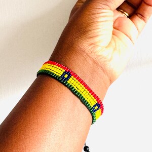 May include: A beaded bracelet featuring horizontal rows of red, yellow, and green beads with a blue square accent containing a yellow center. The bracelet is constructed from small, uniform spherical beads and includes a black cord with a small bead closure.