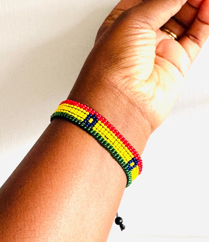 May include: A beaded bracelet featuring horizontal rows of red, yellow, and green beads with a blue square accent containing a yellow center. The bracelet is constructed from small, uniform spherical beads and includes a black cord with a small bead closure.
