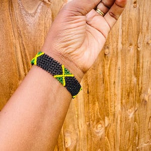 May include: A beaded bracelet featuring a repeating pattern of the Jamaican flag. The design consists of small black, green, and yellow glass beads woven together in a rectangular grid, creating a vibrant and detailed accessory for the wrist.