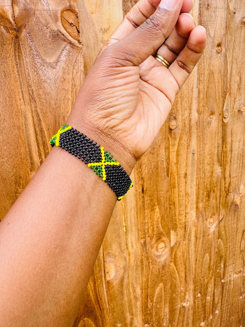 May include: A beaded bracelet featuring a repeating pattern of the Jamaican flag. The design consists of small black, green, and yellow glass beads woven together in a rectangular grid, creating a vibrant and detailed accessory for the wrist.
