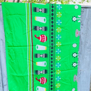 Pu&ograve; includere: Un tessuto verde brillante con un motivo ripetuto di forme geometriche bianche, rosse e nere. Il design include strisce verticali gialle, bianche e rosse. I bordi hanno una frangia.