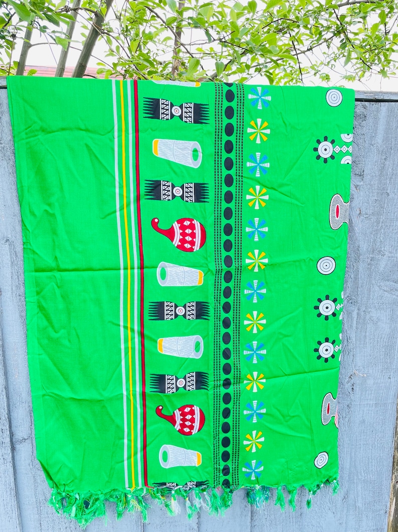 Pu&ograve; includere: Un tessuto verde brillante con un motivo ripetuto di forme geometriche bianche, rosse e nere. Il design include strisce verticali gialle, bianche e rosse. I bordi hanno una frangia.