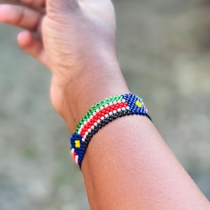 May include: A woven beaded bracelet featuring a repeating pattern of small glass beads in vibrant colors including red, white, green, black, and blue, with small yellow accents arranged in a geometric design around the circumference of the wrist.