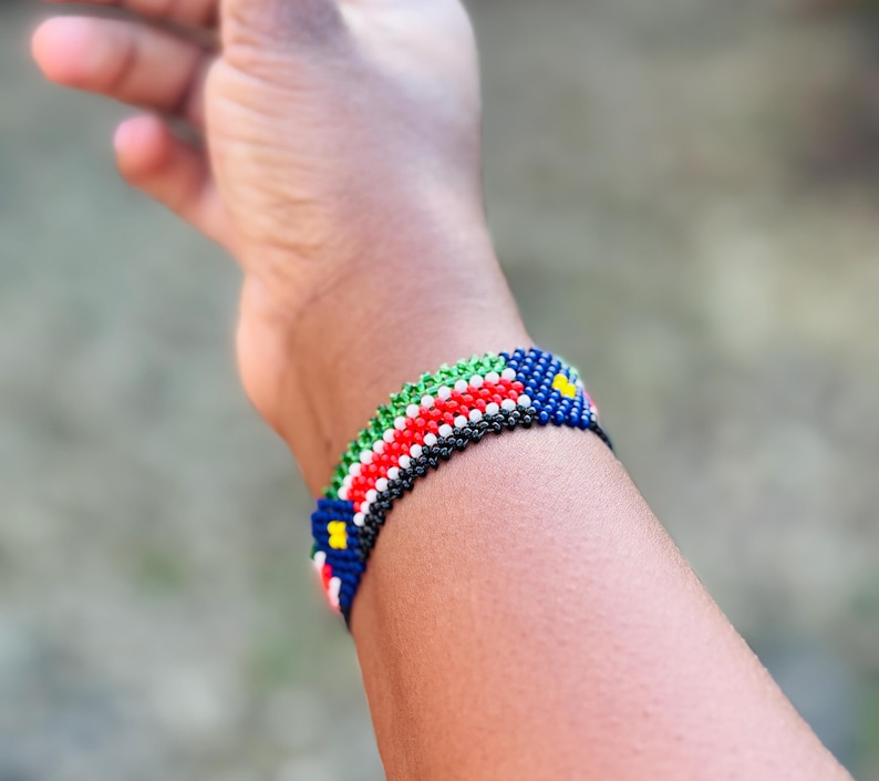 May include: A woven beaded bracelet featuring a repeating pattern of small glass beads in vibrant colors including red, white, green, black, and blue, with small yellow accents arranged in a geometric design around the circumference of the wrist.