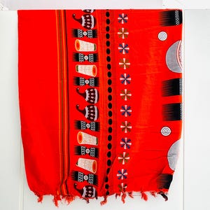 Pu&ograve; includere: Un tessuto rosso vibrante con un motivo dettagliato. Il design include motivi ripetuti di tamburi stilizzati e forme geometriche in nero, bianco e marrone. I bordi sono rifiniti con nappe.