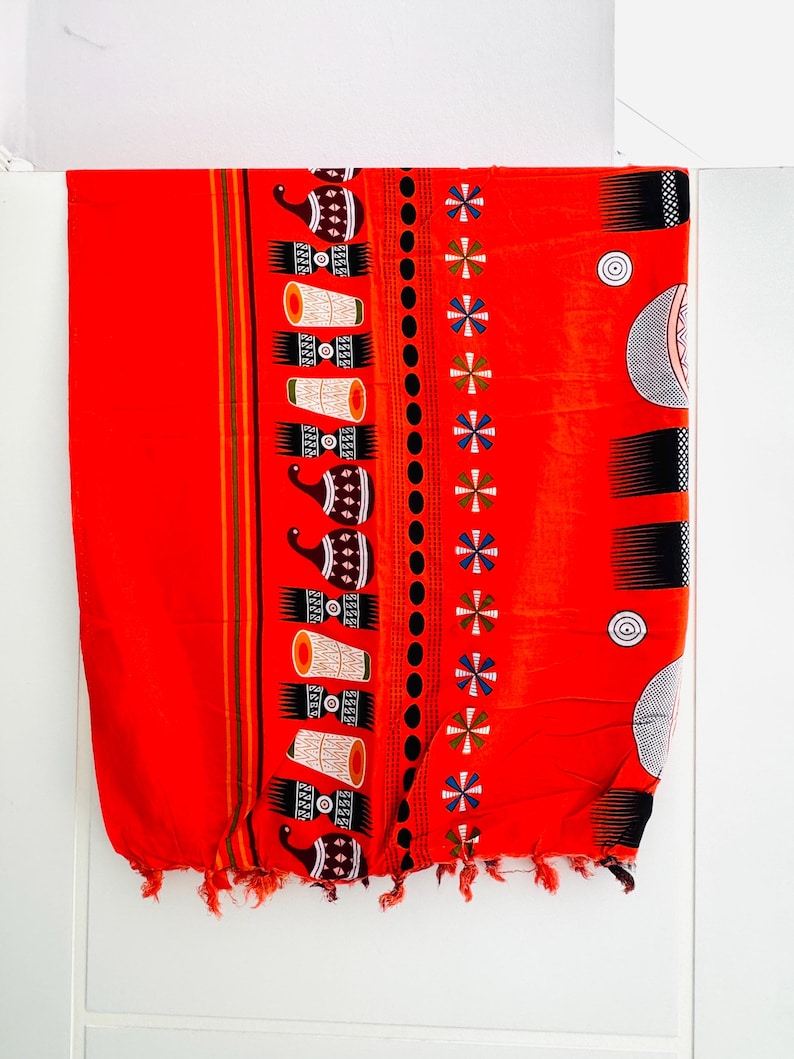 Pu&ograve; includere: Un tessuto rosso vibrante con un motivo dettagliato. Il design include motivi ripetuti di tamburi stilizzati e forme geometriche in nero, bianco e marrone. I bordi sono rifiniti con nappe.