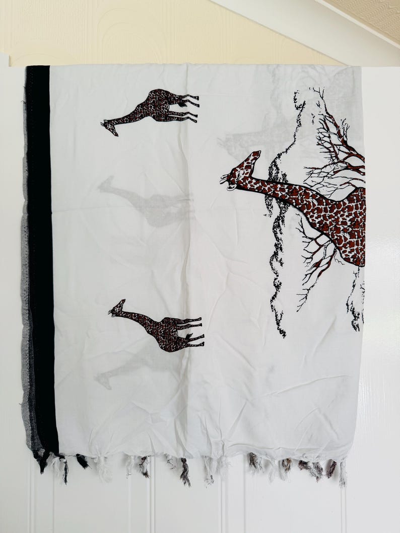 Pu&ograve; includere: Sciarpa bianca con bordo e frange nere. La sciarpa presenta un motivo ripetuto di giraffe marroni con un design maculato. &Egrave; presente anche un disegno simile a un albero sulla sciarpa.