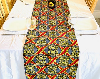 Peut inclure: Un chemin de table bleu, rouge et jaune avec un motif géométrique. Le chemin de table est sur une nappe blanche avec deux assiettes blanches et deux verres à vin.