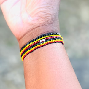 May include: A woven bracelet featuring horizontal rows of small beads in black, red, yellow, and white. The design consists of multiple parallel bands of color, with a small white rectangular accent centered within the black, red, and yellow pattern.