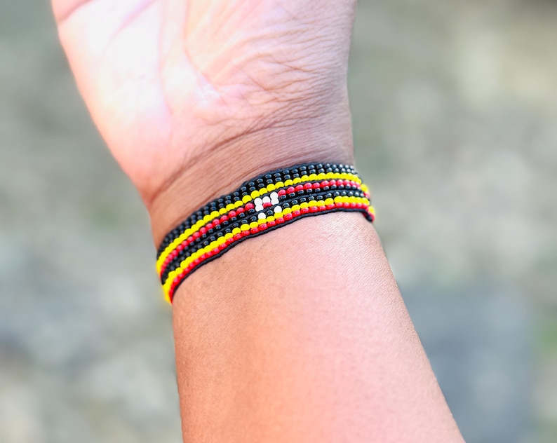 May include: A woven bracelet featuring horizontal rows of small beads in black, red, yellow, and white. The design consists of multiple parallel bands of color, with a small white rectangular accent centered within the black, red, and yellow pattern.