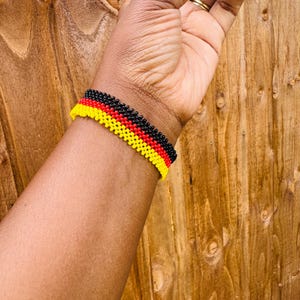 May include: A beaded bracelet featuring three horizontal rows of small, uniform beads in black, red, and yellow. The bracelet is constructed with a woven pattern, creating a textured surface that wraps around the wrist in a consistent, repeating design.
