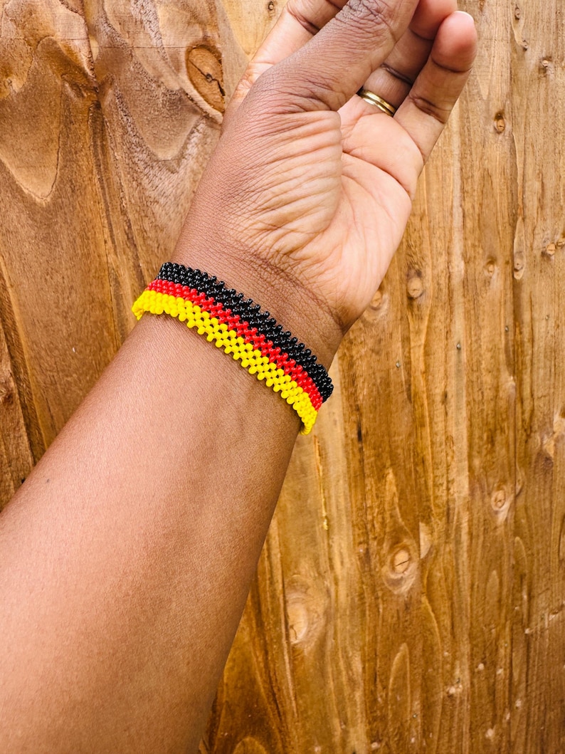 May include: A beaded bracelet featuring three horizontal rows of small, uniform beads in black, red, and yellow. The bracelet is constructed with a woven pattern, creating a textured surface that wraps around the wrist in a consistent, repeating design.