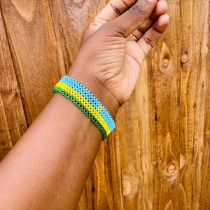 May include: A beaded bracelet featuring horizontal stripes of light blue, yellow, and green glass beads. The bracelet is constructed with a woven pattern and includes a small yellow sun emblem centered within the light blue section of the design.
