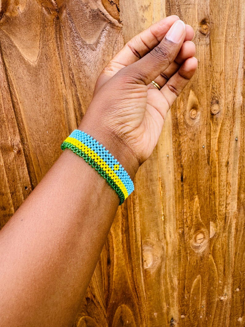 May include: A beaded bracelet featuring horizontal stripes of light blue, yellow, and green glass beads. The bracelet is constructed with a woven pattern and includes a small yellow sun emblem centered within the light blue section of the design.