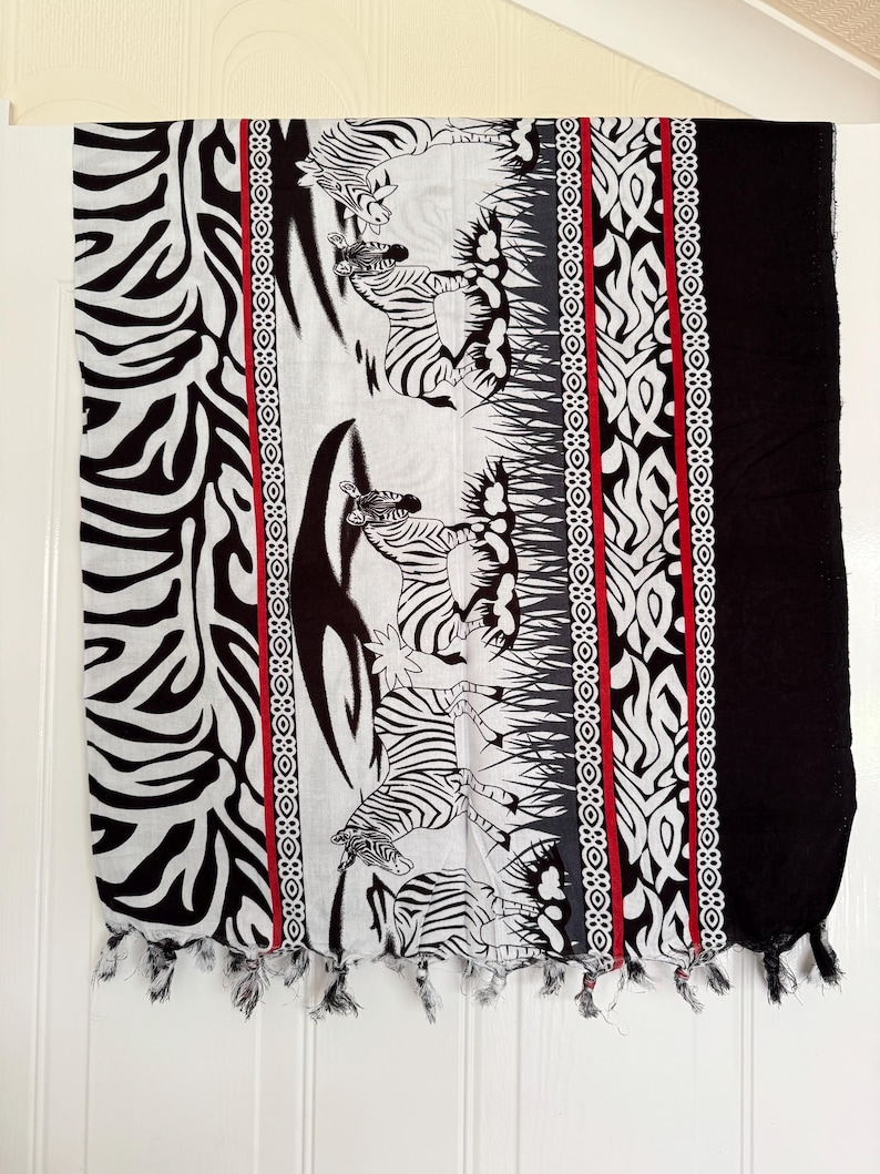 Pu&ograve; includere: Sciarpa bianca e nera con un motivo zebrato. La sciarpa presenta un pannello centrale con illustrazioni di zebre, bordato da motivi decorativi e una striscia rossa. Le nappe adornano il bordo inferiore.