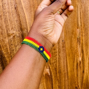 May include: A beaded bracelet featuring horizontal stripes of red, yellow, and green glass beads. The center of the bracelet displays a circular motif with a dark blue background, a yellow four-pointed star shape, and a green border.